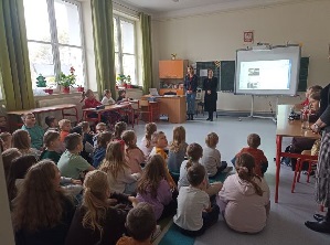 Zdjęcie przedstawiające siedzące tyłem dzieci w klasie, słuchajace prowadzącej.