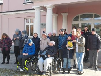 Zdjęcie przedstawia grupę ludzi przed budynkiem. Dwie osoby na wózkach inwalidzkich. Kobieta i dziecko trzymają kartkę ze zdjęciem żółtych krokusów.